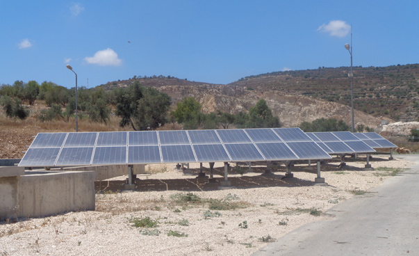 Eine Anordnung von Solarmodulen steht auf einem trockenen Boden. Im Hintergrund sind bei blauem Himmel Hügel zu erkennen.